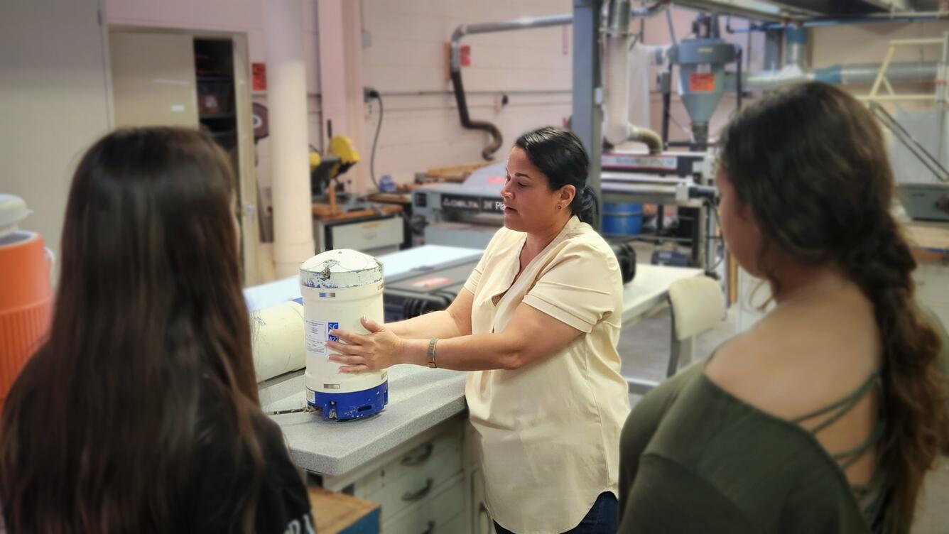 a scientist points to a cylindrical object in front of two young women in a laboratory