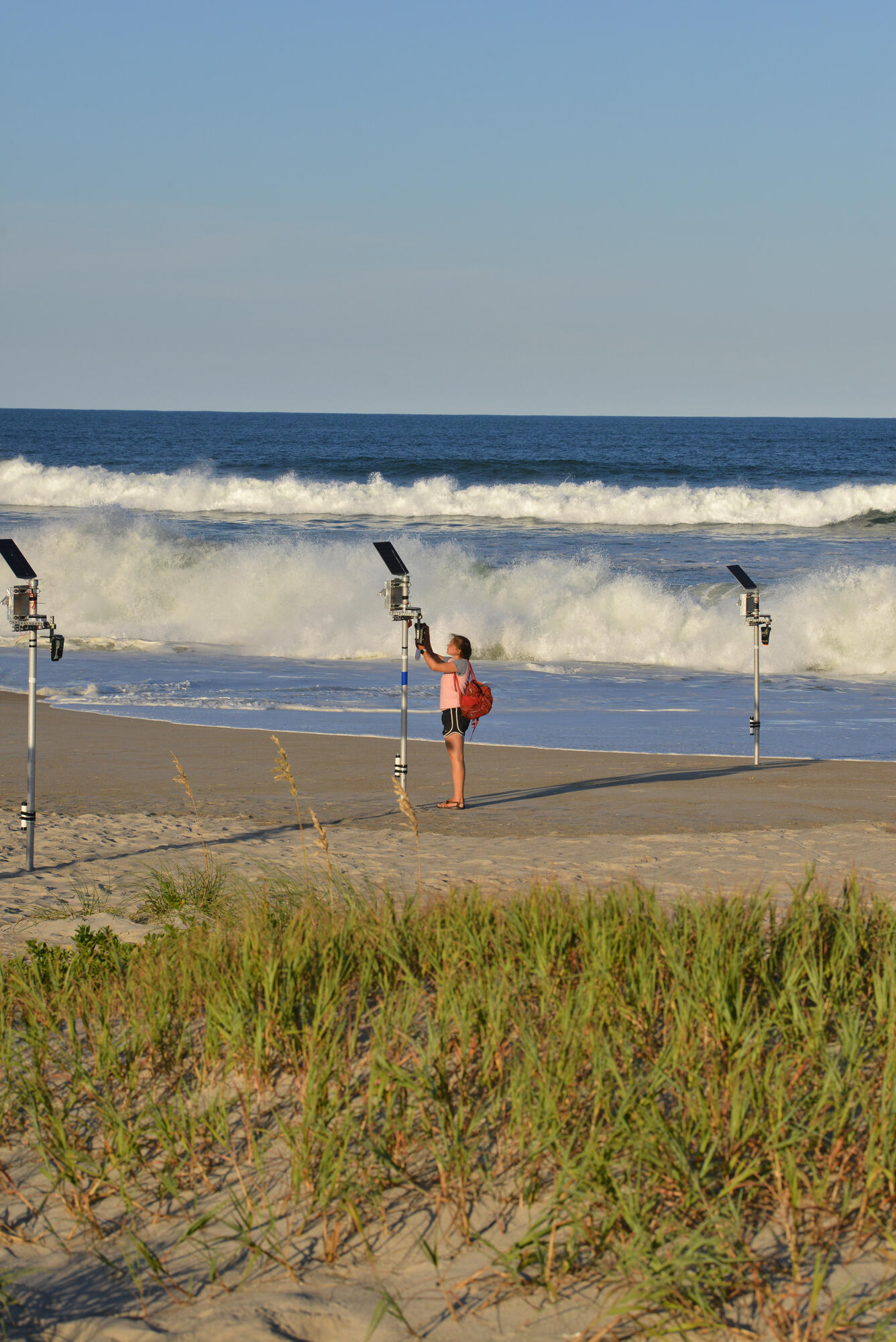 dune grass, sandy beach with three metal pole instruments leading to ocean, person working on one, ocean and sky