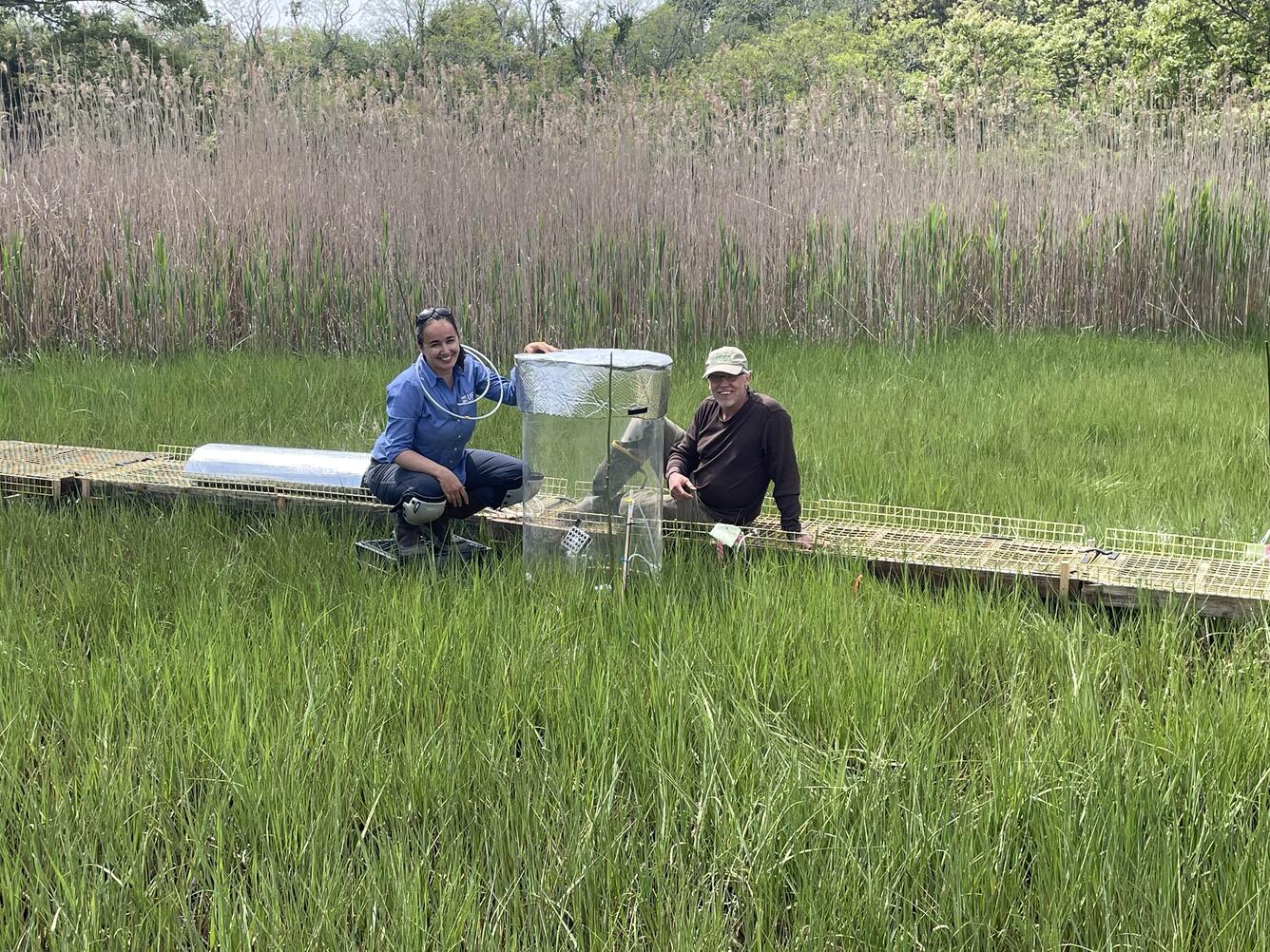 Two people in the middle of an estuary smiling at the camera while sitting on a ramp working with carbon flux equipment