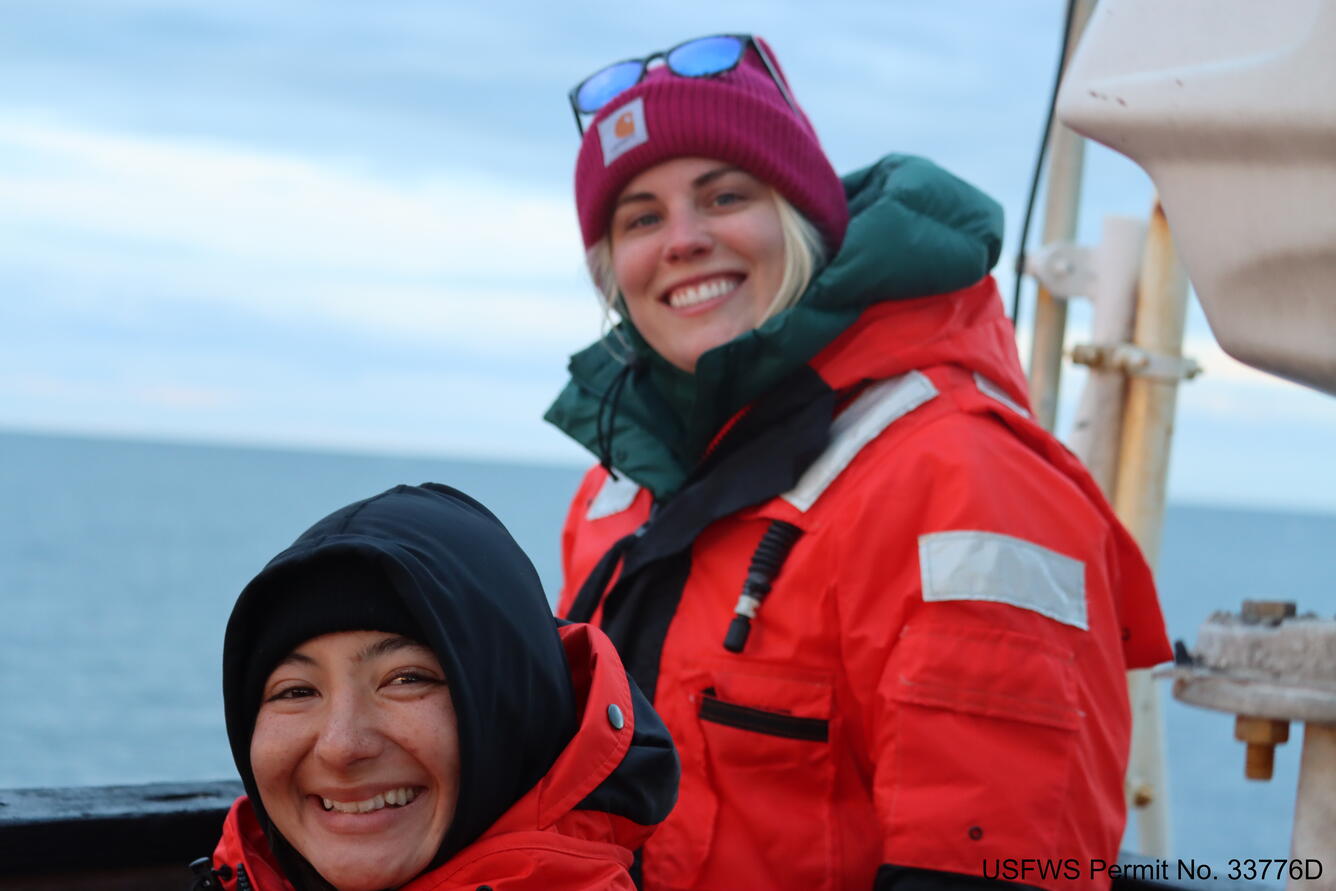Amanda and Ami are on top of the wheelhouse of the ship. Both women are wearing orange flotation survival suites and caps.