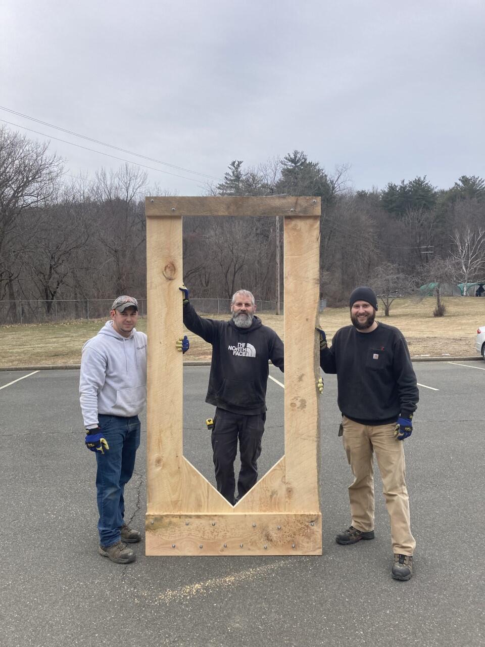 3 men hold a 9 foot tall wood frame upright in a parking lot