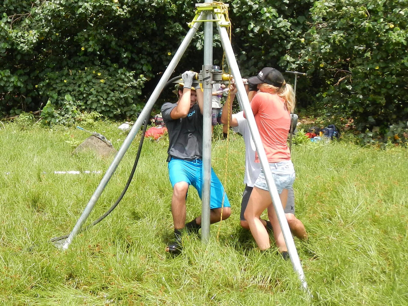 Three people pull hard on handles on a coring device stabilized by a tripod frame, to push and twist it into a grassy marsh.
