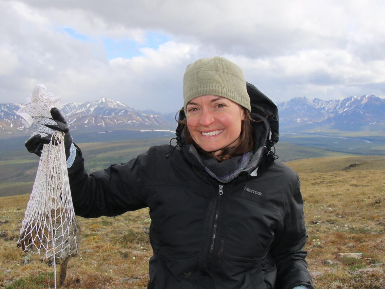 Woman in a coat standing on a plain with mountains in the background. She is holding something in a net.
