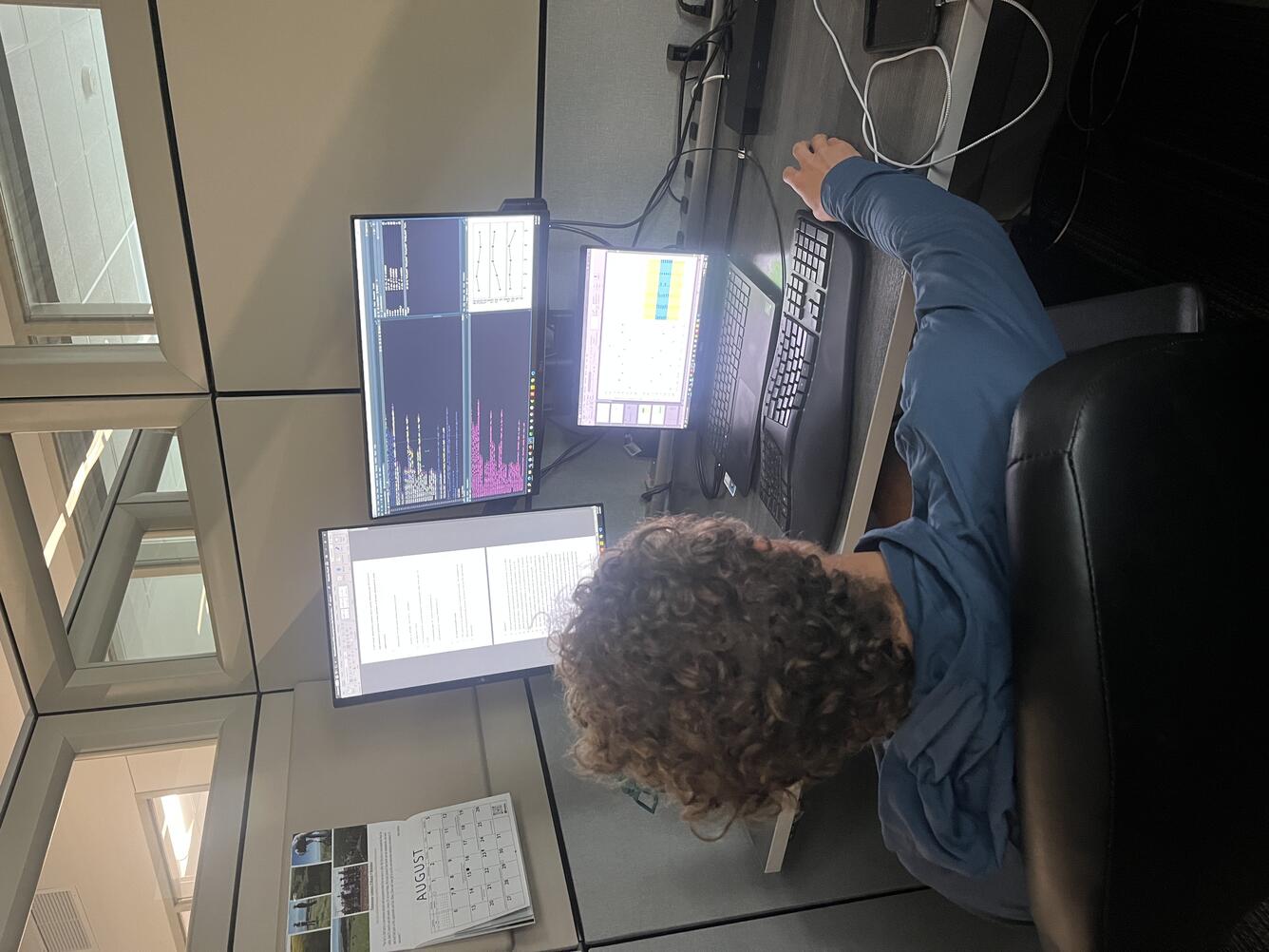 a person sitting at a desk facing away from the camera looking at 3 computer monitors