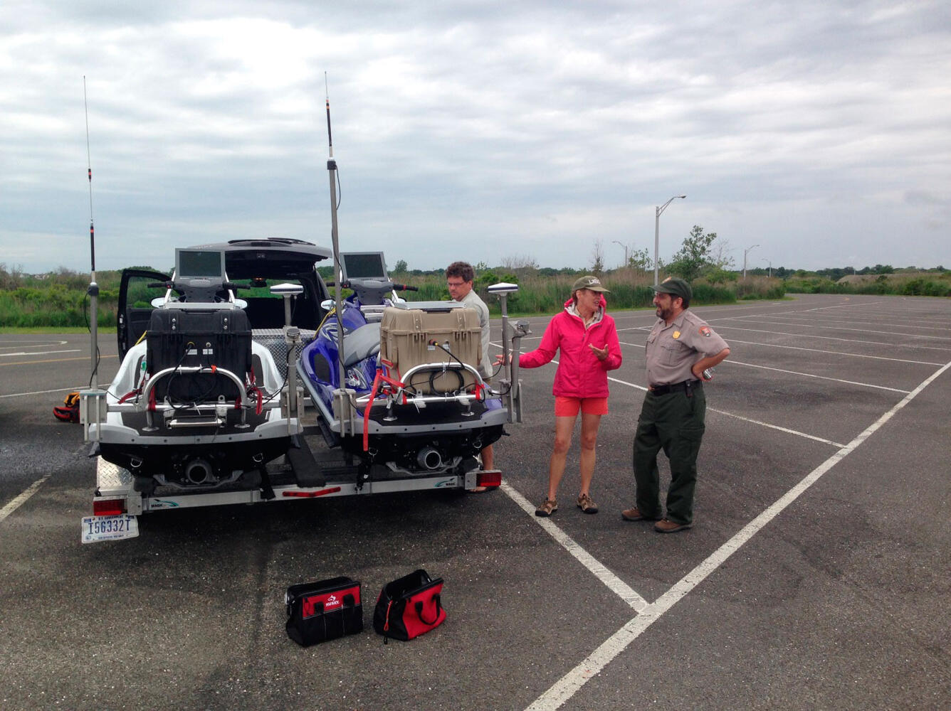 Three people standing in a parking lot near a trailer with two jet skis on it.