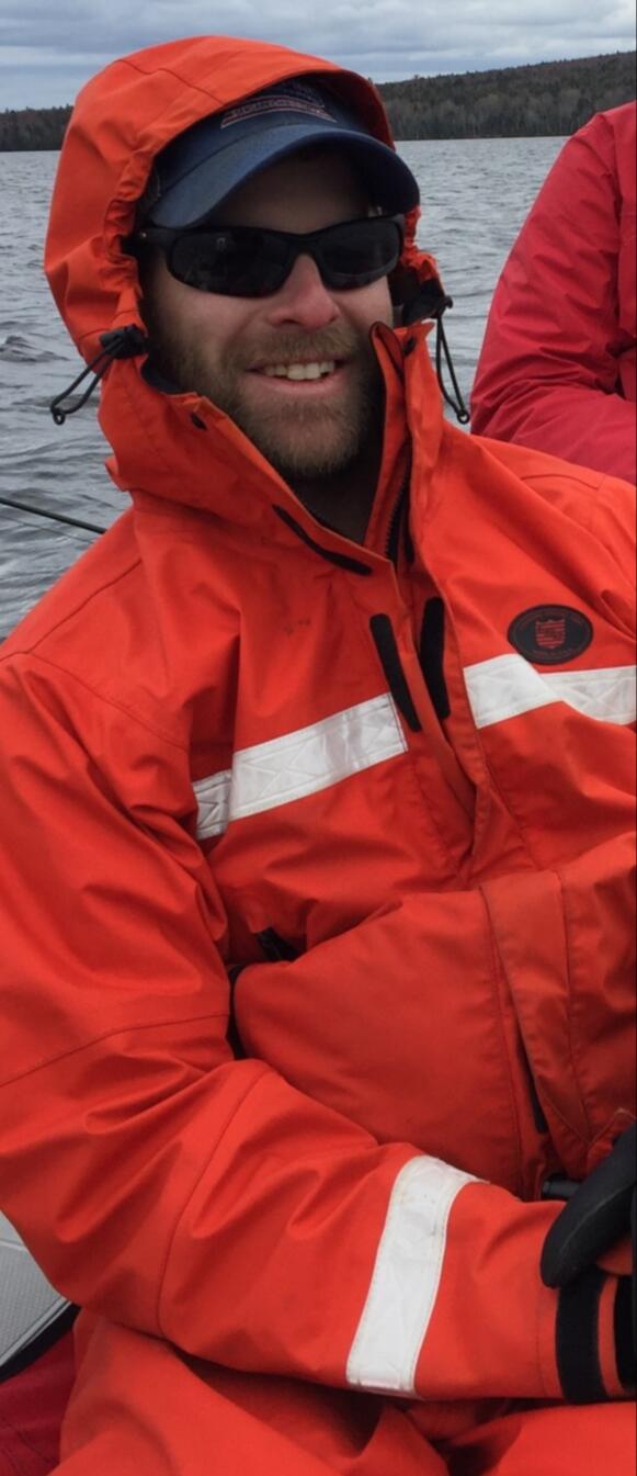 photograph of a bearded man wearing sunglasses and an orange raincoat in a boat on a lake