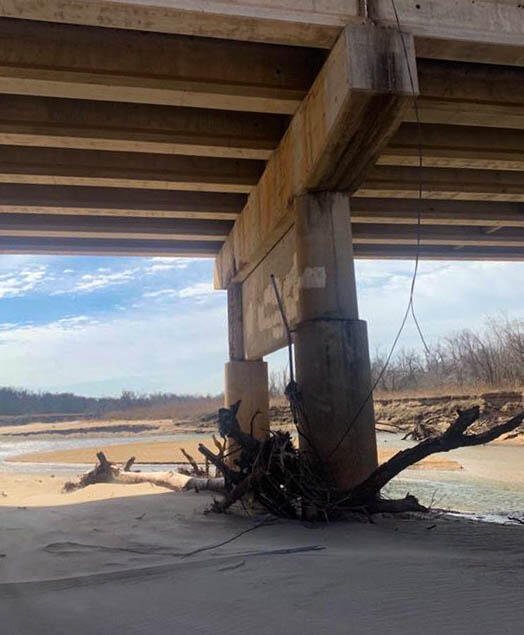Wide sandy channel under a bridge, with a narrow meandering river and several large pieces of tree debris against the pilings