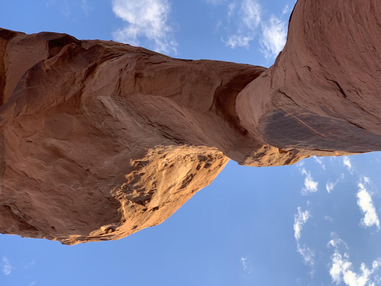 This image shows the under side of a red sandstone arch against a bright blue sy with a few white clouds.