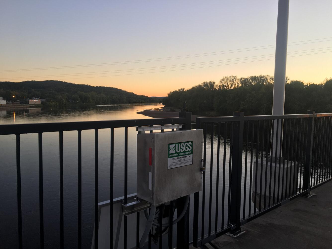 Streamgage mounted to bridge, view facing downstream river at dusk