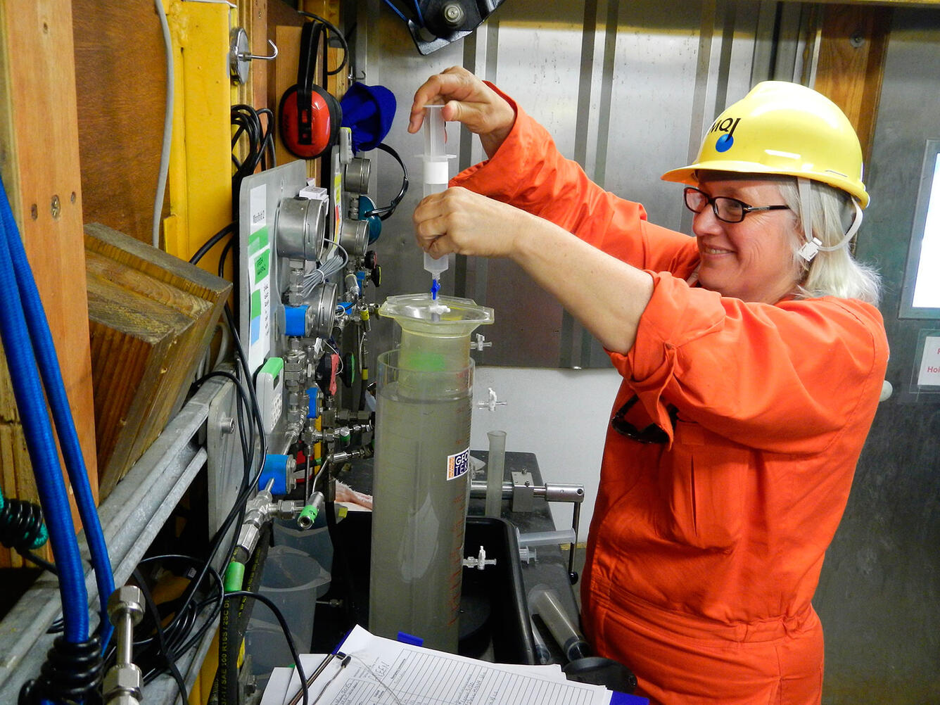 A woman wearing an orange jumpsuit and hard hat draws a sample with a syringe from a tall cylinder filled with murky water.