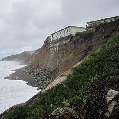 Photo of a high cliff crumbling into the sea with buildings right on top of it.