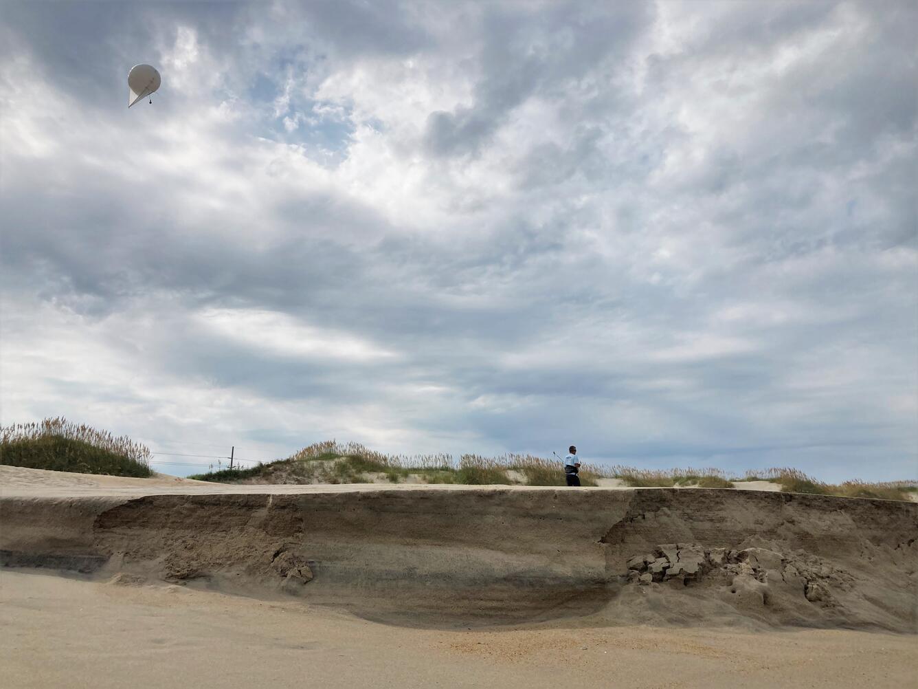 A person walks along a sandy scarped dune near the waves under a cloudy sky, pulling along a white balloon-shaped kite