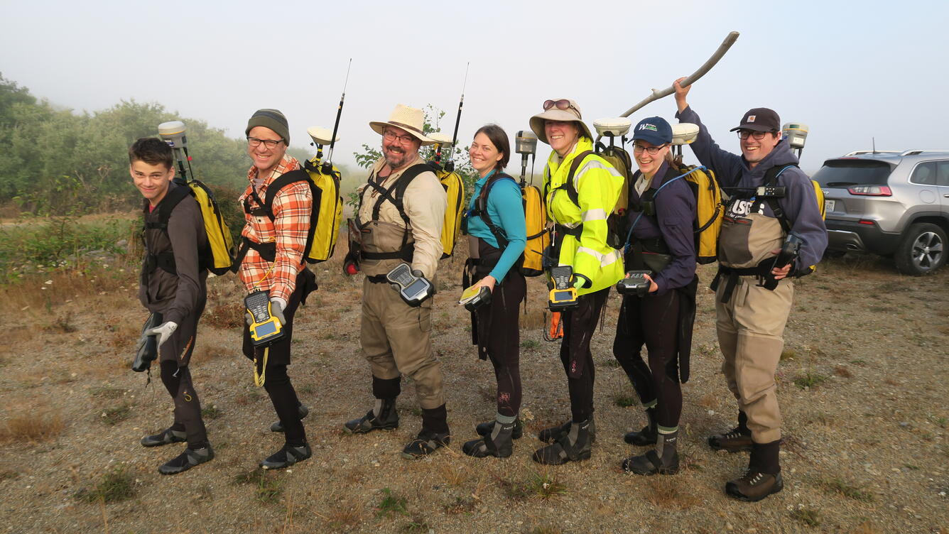People are wearing wet suits and waders and are holding hand-held computers and backpacks with equipment in them, smiling.