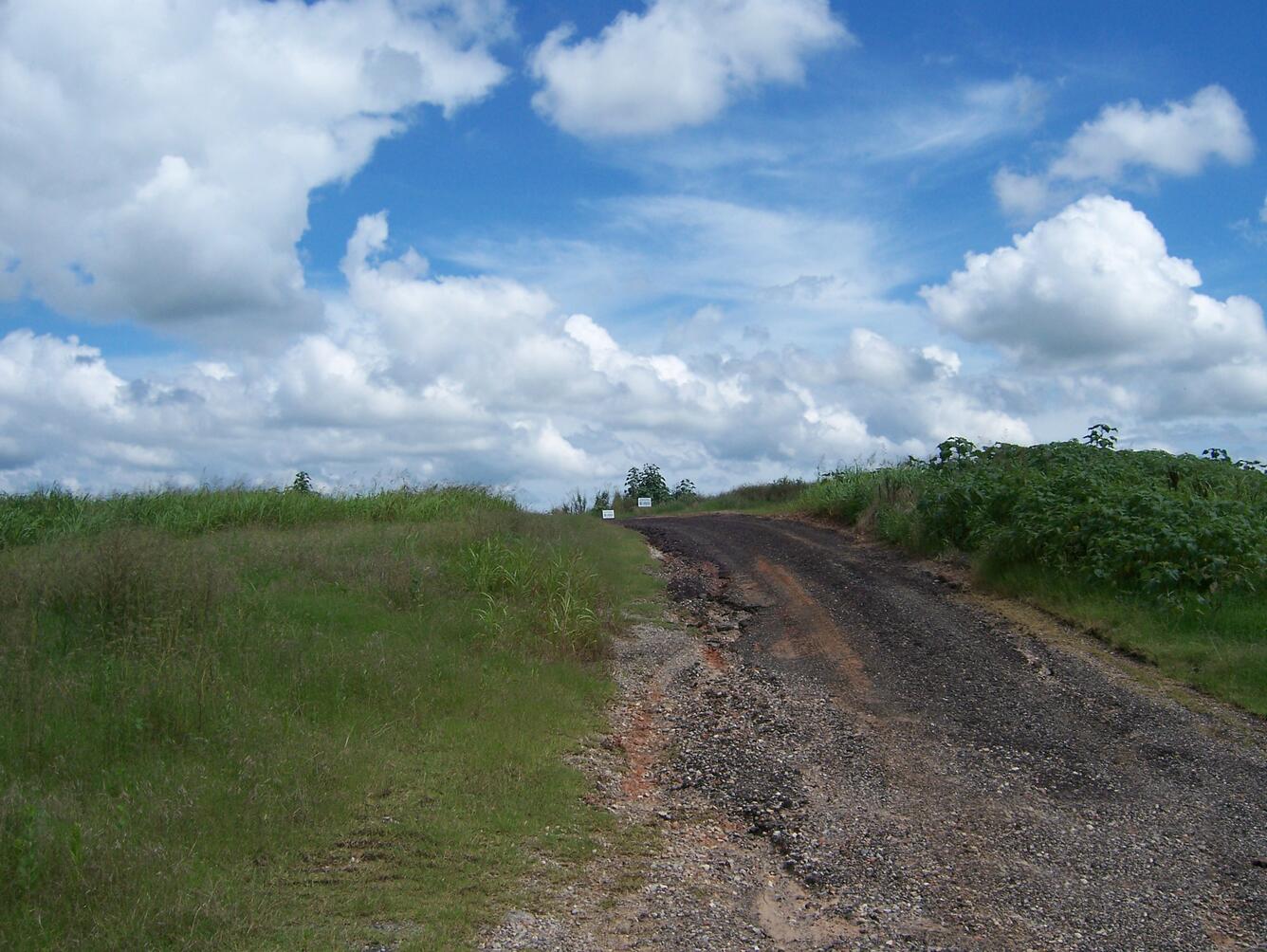 Photo of a rough asphalt road laid on the side of a landfill. Two USGS well signs are in the distance.