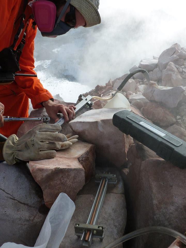 A scientist leans over a pile of rocks, wearing a gas mask and taking measurements from an instrument inserted into the ground
