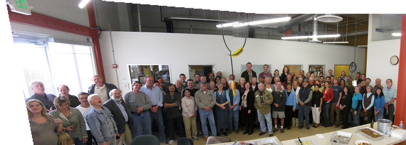 A large group of people stand together for a team portrait in a space that looks like a warehouse with offices.
