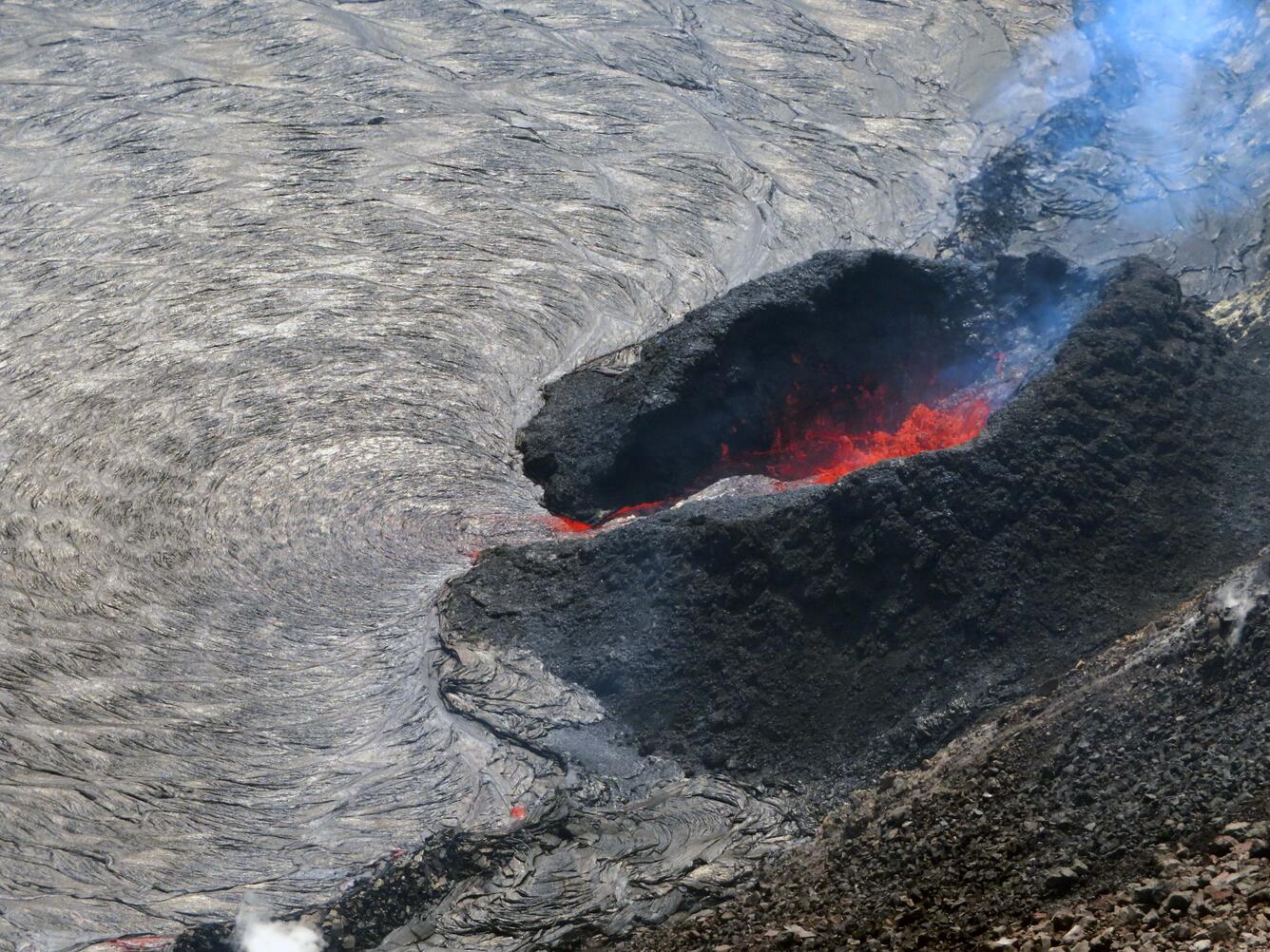 Color photo of a lava fountain at the edge of a lava lake with a dark spatter cone around it