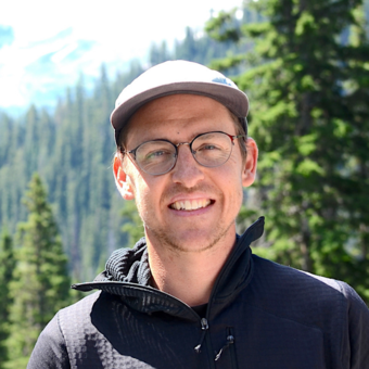 Headshot of Tyler Hoecker wearing a hat and glasses and standing in front of a forested mountain.