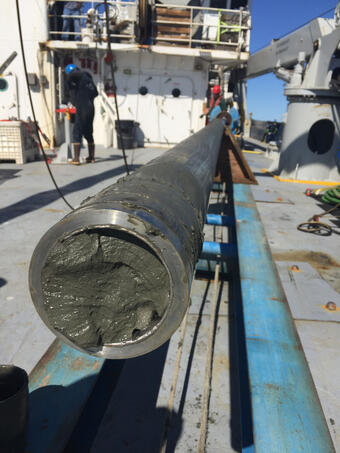 Photo looks at the very end of a metal coring device with mud in it, sitting on the deck of a ship in its metal launch.