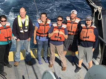 A group of 6 people wearing lifejackets stand on a ship smiling up at the camera person.